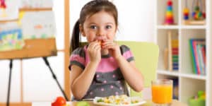 Young girl eating a meal at a table in a colorful classroom setting.