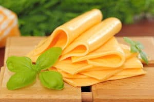 Folded slices of unwrapped yellow cheese stacked on a wooden board with fresh basil.